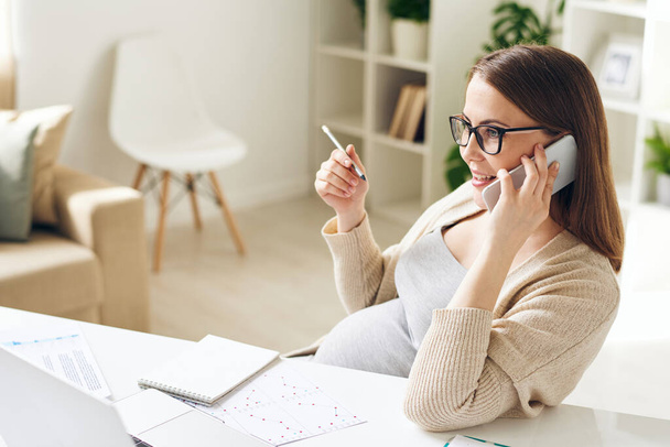 Confident young pregnant businesswoman in glasses sitting at desk with papers and answering client by phone in office - Foto, afbeelding