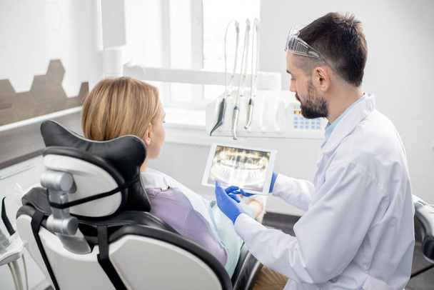 Νεαρός άνδρας οδοντίατρος με γάντια και λευκή ρόμπα που εξηγεί στην ασθενή του όλα τα προβλήματα με τα δόντια της, ενώ δείχνει ακτινογραφία στο δισκίο - Φωτογραφία, εικόνα