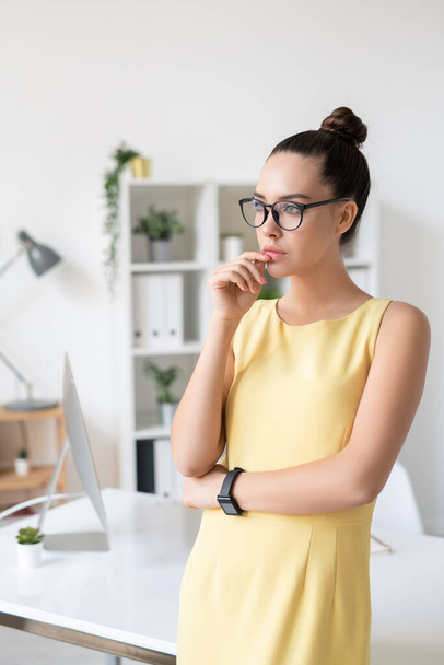 Joven empresaria emprendedora pensativa en vestido amarillo y gafas de pie con la mano cerca de los labios en la oficina moderna
 - Foto, imagen