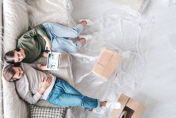 Přehled mladého láskyplného páru s tabletem sedícím na novém gauči a prohlížejícím si inzeráty interiérů nebo prodávajícím nemovitosti - Fotografie, Obrázek