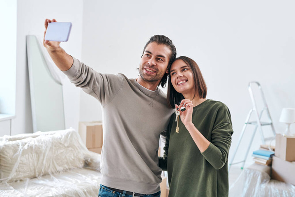 スマートフォンを持つ若い男は自分の幸せな妻と一緒に自分の新しい家やフラットキーを保持しながら、リビングルームに立って - 写真・画像