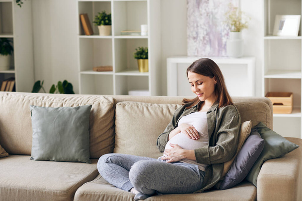 Calmo atraente jovem grávida sentada com pernas cruzadas no sofá e abraçando sua barriga
 - Foto, Imagem