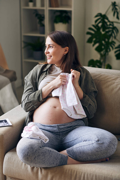 Positive pretty young pregnant woman sitting with babys bodysuit on sofa and dreaming about first meeting with baby - Photo, Image