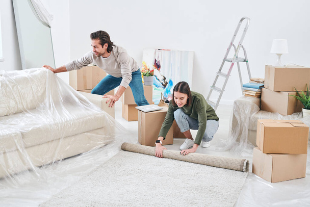 Mujer joven rodando alfombra en el suelo, mientras que su marido poner celofán en el sofá antes de la reparación de su nueva casa o piso
 - Foto, Imagen