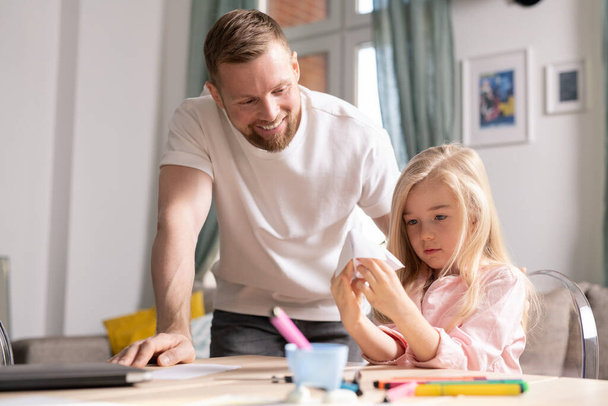 Schattig klein meisje kijkt naar papier speelgoed in haar handen terwijl ze aan tafel zit en haar lachende vader staat in de buurt en buigt over haar heen - Foto, afbeelding