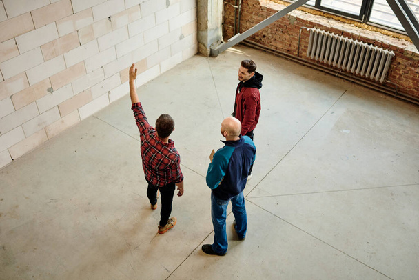 Группа молодых строителей обсуждает качество материалов, в то время как один из них указывает на стену незавершенного здания на стартапе
 - Фото, изображение