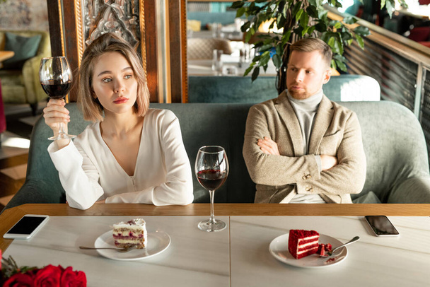 Jolie femme blonde avec un verre de vin rouge regardant par la fenêtre alors qu'elle était assise à table dans un restaurant avec un petit ami ennuyeux près de chez elle
 - Photo, image