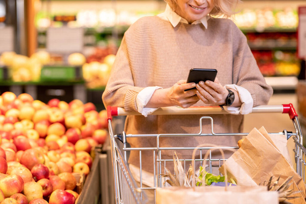 Ώριμη γυναίκα αγοραστής σε casual πουλόβερ κύλιση μέσω της λίστας αγορών στο smartphone, ενώ πιέζει καλάθι με χάρτινες σακούλες στο σούπερ μάρκετ - Φωτογραφία, εικόνα