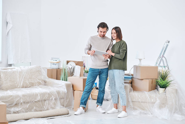 Młody czuły mężczyzna i kobieta w stroju casualwear patrząc na cyfrowy wyświetlacz tabletów i omawiając pomysły online dla ich nowego mieszkania lub domu - Zdjęcie, obraz