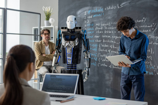 Подросток Клевер с ноутбуком стоит у доски перед классом и проводит презентацию способностей робота и его возможностей - Фото, изображение