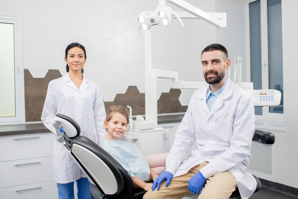 Dwóch młodych profesjonalnych dentystów w rękawiczkach i białych płaszczach i uroczy mały pacjent w fotelu patrzący na ciebie w klinikach dentystycznych. - Zdjęcie, obraz