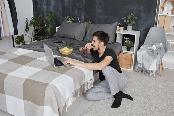Hombre joven en ropa de casa sentado en el suelo en el dormitorio y el uso de la computadora portátil mientras come patatas fritas durante el aislamiento
 - Foto, Imagen