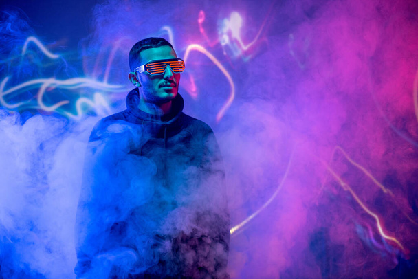 Serious cool guy in neon goggles and sweatshirt standing in blue and pink smoked room - Фото, изображение