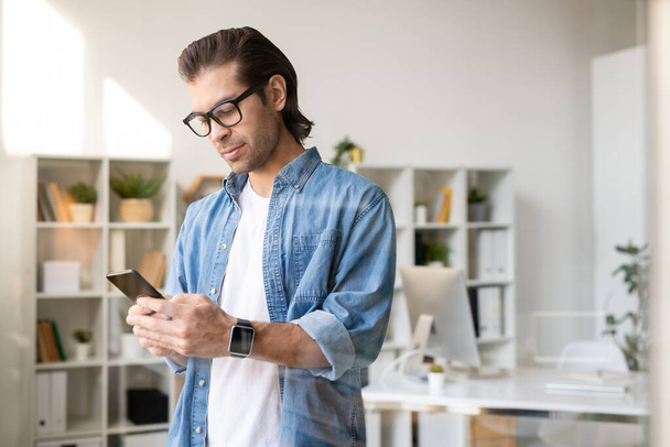 Serious busy young brunette man in glasses checking phone message in own office - Photo, image