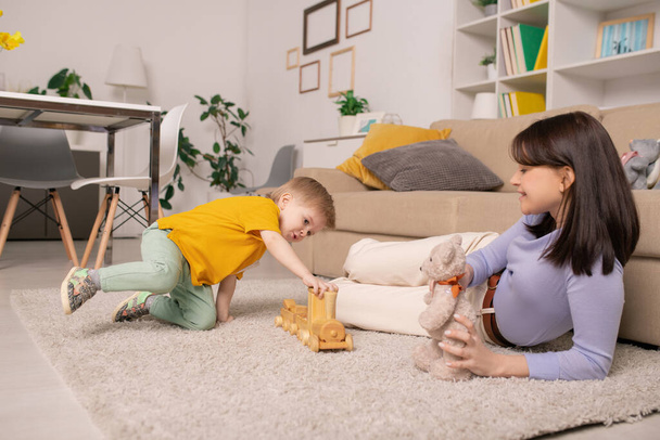 Feliz joven madre acostada en una alfombra suave en casa y sosteniendo un oso de juguete mientras su hijo juega con un tren de juguete
 - Foto, Imagen