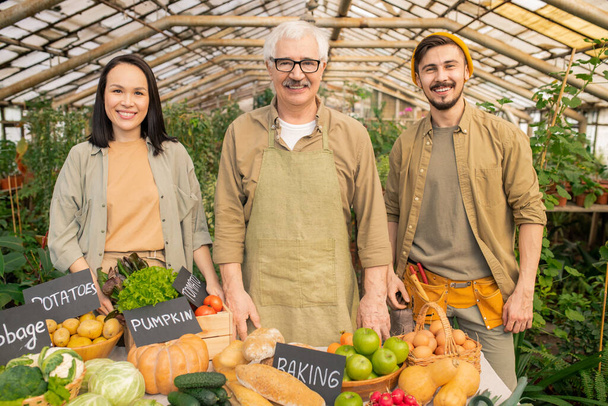 Retrato de agricultor senior positivo en delantal vendiendo alimentos orgánicos con jóvenes asistentes en el mercado orgánico
 - Foto, imagen