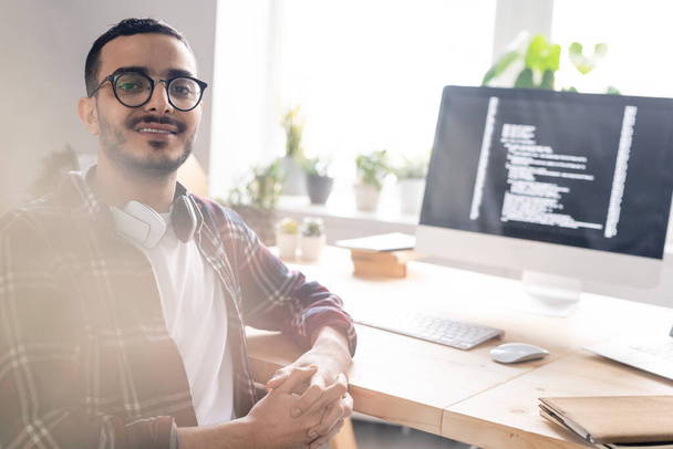 Retrato de un joven codificador positivo de Oriente Medio en gafas sentadas con las manos apretadas en el escritorio con una computadora potente
 - Foto, imagen