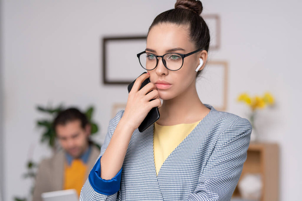Pensive young businesswoman in blue checkered jacket wearing wireless ear buds holding phone near face while thinking of project - Fotoğraf, Görsel