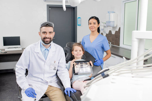 Szczęśliwy młody dentysta w białym płaszczu, jego mały pacjent i asystent w mundurze patrząc na ciebie w miejscu pracy we współczesnych klinikach stomatologicznych - Zdjęcie, obraz
