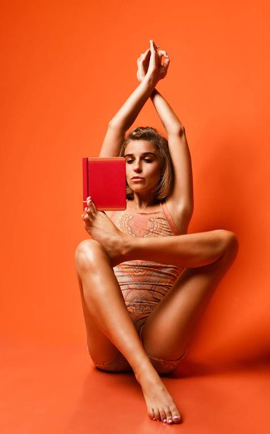 young fitness woman in sportswear doing yoga, stretching her leg, reading a book on studio backdrop orange background. athletic girl exercise, training, workout, stretching, relaxation - Photo, image