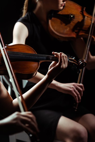 partial view of two female professional musicians playing on violins on dark stage  - Foto, imagen