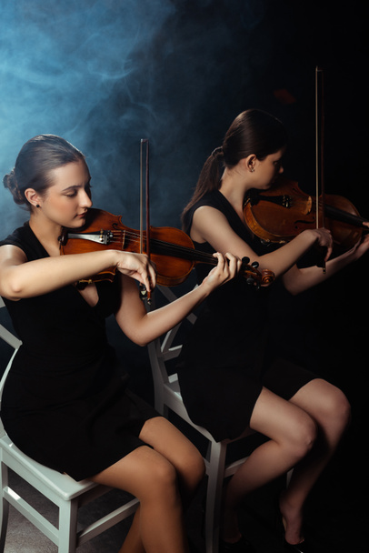 attractive professional musicians playing on violins on dark stage with smoke - Photo, Image