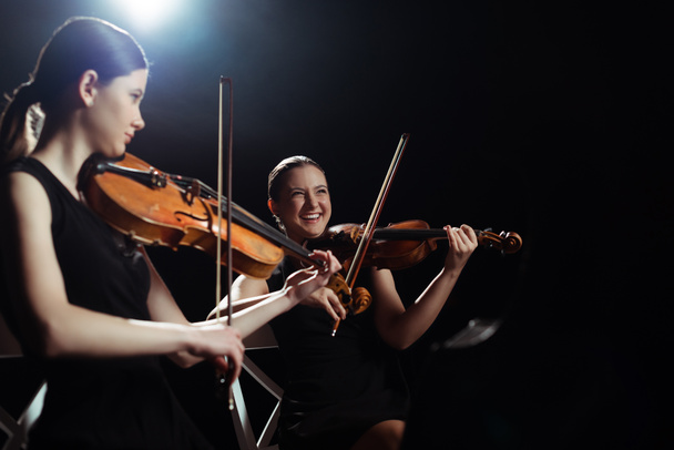 laughing female musicians playing on violins on dark stage with back light - Photo, Image