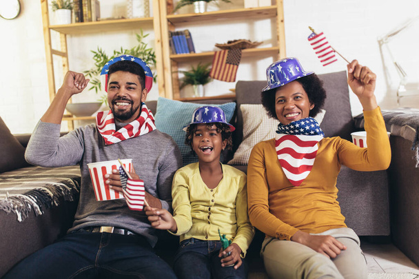 Счастливая афроамериканская семья из трех человек смотрит телевизор и весело проводит спортивные игры на диване дома
 - Фото, изображение