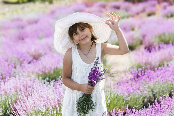 Feliz linda niña está en un campo de lavanda está usando vestido blanco sosteniendo ramo de flores púrpura
. - Foto, Imagen