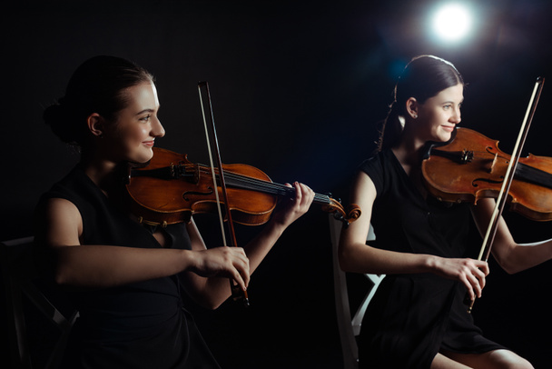 happy attractive musicians playing on violins on dark stage with back light - Photo, Image