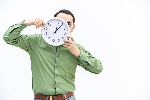 Young man in green is waiting, holding clock in front of his face over white background. - Foto, immagini