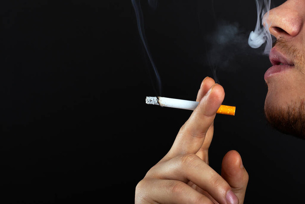 Ein junger Mann hält eine Zigarette in der Hand und atmet vor dunklem Hintergrund Rauch aus. Schaden durch Nikotin- und Tabakkonsum   - Foto, Bild