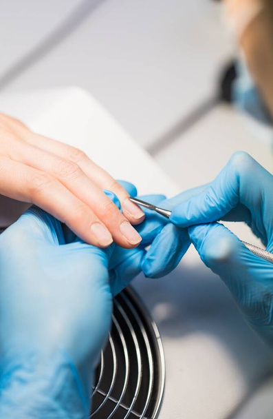 Manicurist in blue hygienic gloves do manicure of nails in nail salon. Woman using nail file - Photo, Image