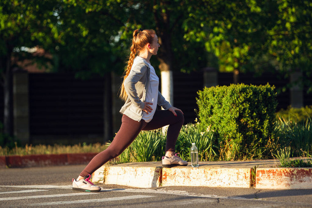 Young female runner, athlete is stretching before jogging in the city street in sunshine. Beautiful caucasian woman training, listening to music - Foto, immagini