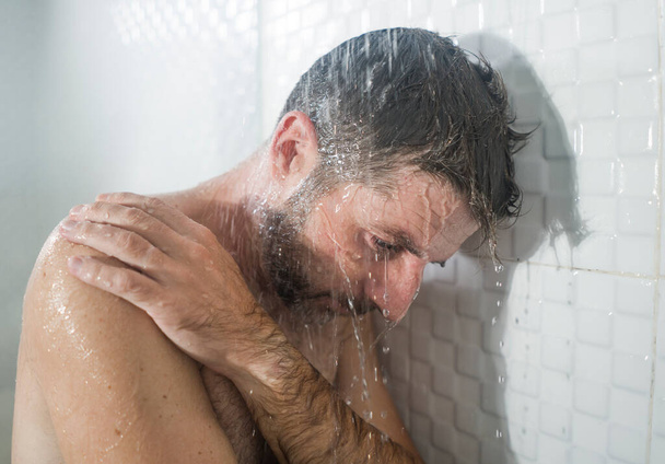 home bathroom dramatic portrait of young attractive sad and depressed man in the shower leaning against the wall thoughtful feeling overwhelmed by life problem  - 写真・画像