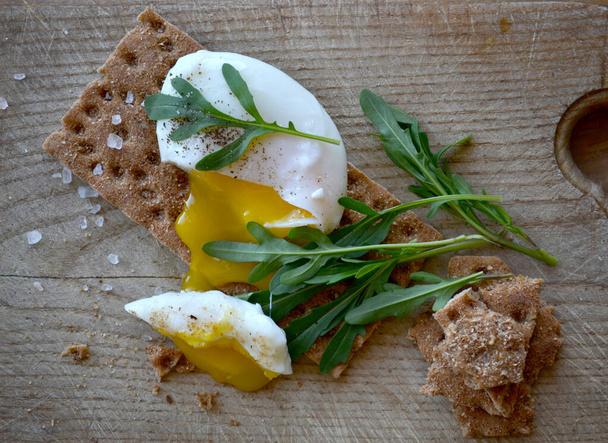 Pochiertes Ei auf Vollkornbrot mit Rucola auf einem Holztisch. Leckeres und gesundes Frühstück. - Foto, Bild