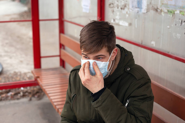 Молодой человек сидит на автобусной станции в одноразовой медицинской маске крупным планом. карантин из-за вирусных респираторных заболеваний covid-19
.  - Фото, изображение