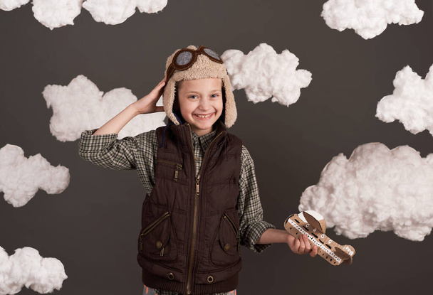 ένα κορίτσι παίζει με ένα αεροπλάνο από χαρτόνι και ονειρεύεται να γίνει πιλότος, ντυμένος με ένα ρετρό στυλ σακάκι και κράνος με γυαλιά, σύννεφα από μαλλί βαμβάκι, γκρι φόντο, χρωματισμένο σε καφέ - Φωτογραφία, εικόνα