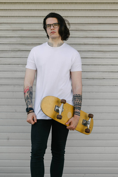 Hipster handsome male model wearing white blank t-shirt with space for your logo or design in casual urban style. He is holding a skateboard - Foto, immagini