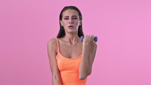Young tired displeased fitness woman isolated over pink wall background make exercises with dumbbells - Кадры, видео