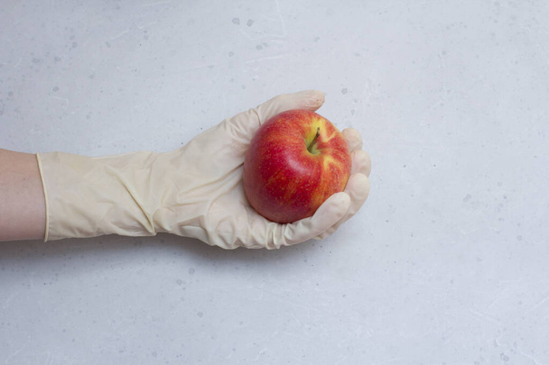 Czerwone jabłko leży pod ręką w białej gumowej rękawicy jednorazowego użytku, na jasnoszarym i białym, nowoczesnym tle betonowym. Ręka w gumowej rękawiczce trzyma owoc jabłka. Koncepcja epidemii koronawirusów. - Zdjęcie, obraz