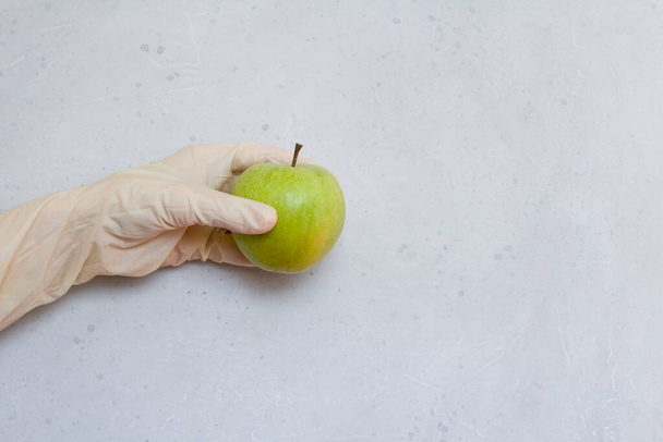 Una mano en un guante de goma sostiene una fruta de manzana verde. Manzana verde se encuentra en la mano en guante de látex de goma desechable blanco, sobre fondo de hormigón moderno gris claro y blanco. Concepto epidémico de Coronavirus
. - Foto, Imagen