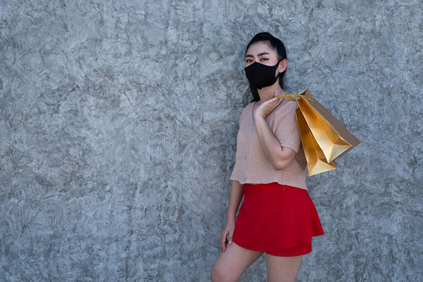 Portrait of a beautiful young Asia woman wearing a face mask with holding shopping bags at concrete wall background, Women wear pink clothes and red skirt looking at the camera, shopping sale concept - Photo, Image