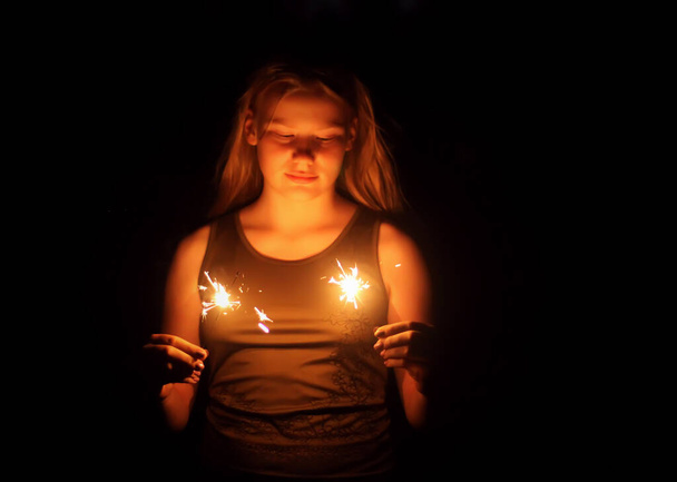 Teenager girl holding a burning sparklers in her hands on night darkness background. - Photo, Image