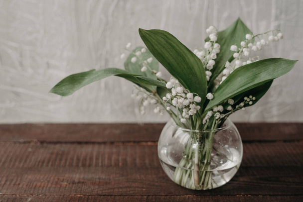 Lily of the valley flowers in glass vase, cement background, selective focus.Still life with a bouquet of white lilies of the valley in a glass vase on the table. - Photo, Image
