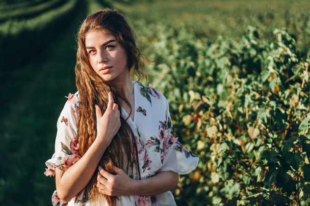 beautiful woman with long curly hair and freckles face on currant field background. Girl in a light dress walks in the summer sunny day. - Foto, afbeelding
