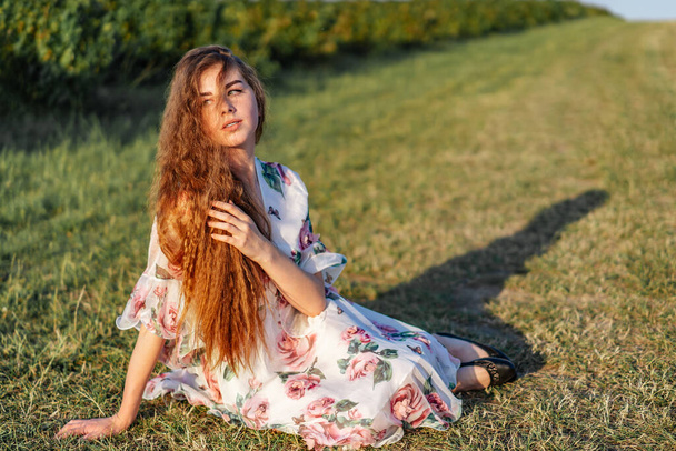 Full length portrait of beautiful woman with long curly hair on currant field background. Girl in a light dress sits on the grass in sunny day. - Foto, Bild