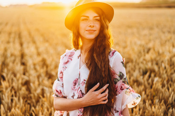 Portrait of a beautiful young woman with curly hair and freckles face. Woman in dress and hat posing in wheat field at sunset and looking at camera. - Valokuva, kuva