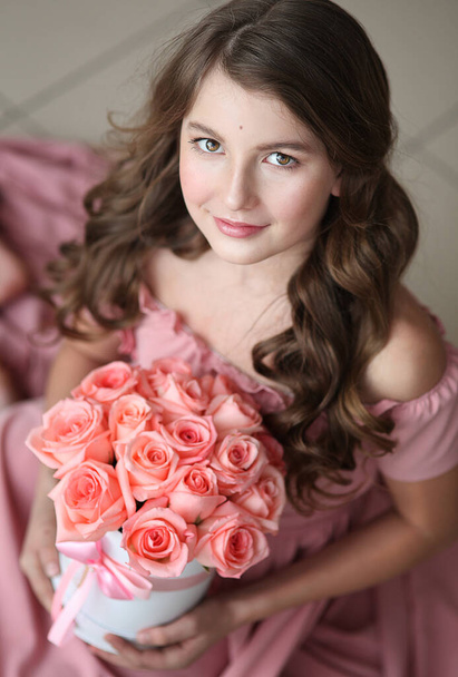 Portrét mladé dívky s dlouhými krásnými kudrnatými vlasy v růžových dlouhých šatech s kyticí růžových růží v krabici, sedí a dívá se přímo do kamery - Fotografie, Obrázek