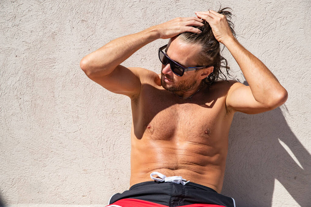 The handsome brutal man in sunglasses with a long hair and naked torso sits near the pool, collects hair in a tail, a sports suntanned body, sunglasses with a blue frame, red blue swimming shorts - Фото, изображение